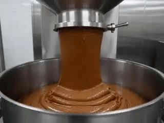 Thick brown industrial paste pouring from a steel factory pipe into a vat